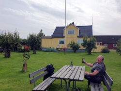 Rasenfläche, im Hintergrund ein gelbes Holzhaus. Eine Frau hebt eine Bierdose zum Zuprosten.